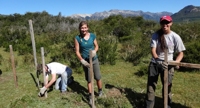 Three people work on fence posts during a service project with outward bound. There are mountains in the background. 